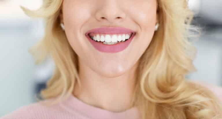 Unrecognizable blonde lady showing beautiful white teeth after treatment, sitting at dental chair, cropped, panorama. Woman attending modern dental clinic, having teeth whitening treatment